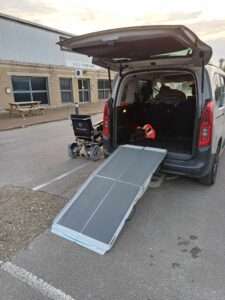 the back of a car with the boot open, ramps leading into with an powered wheelchair stationed next to the car