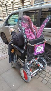 A overweight woman in a power wheelchair dressed up as a ghost/fairy type Pokemon trainer. Sporting giant purple butterfly wings on her back, a sign on the back of the chair tells everyone she is a pokemon trainer. The wheels of the chair are covered with red and white discs to make them look like pokeballs.