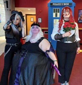 A trio of plucky adventurers posing in front of the TARDIS. Hero on the left is wearing a black hat, black and white wig and a black outfit. The Hero on the right is wearing a long red curly wig, white shirt and black trousers and is holding a bunch of roses. The hero in the middle, sat in a wheelchair, sporting a white wig, a purple painted face and wearing a black dress with black and purple tentacles at her side.