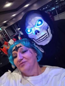 A skeleton with glowing blue eyes, looking over the shoulder of a blue haired women with jewels and glitter on her face. You can just see the snakes hiding in her hair.