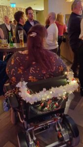 The back view of an overweight woman, sat in a power wheelchair, she is wearing a Christmas print dress and the back of the chair has fairy lights, tinsel and a string of bells attached.