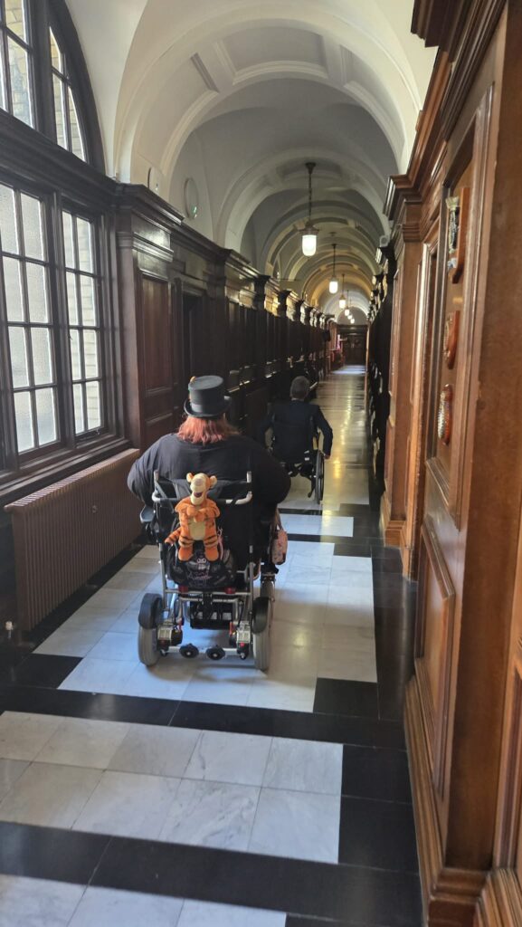 A long stately corridor with high arched ceiling, two figures in wheelchairs look like they are racing each other into the distance.