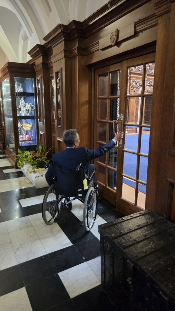 A man in a wheelchair, reaching up to open a large glass panelled door