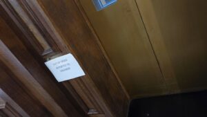 A white piece of paper with black writing on it saying "Out of Order, reported to engineer" taped to the lift call button