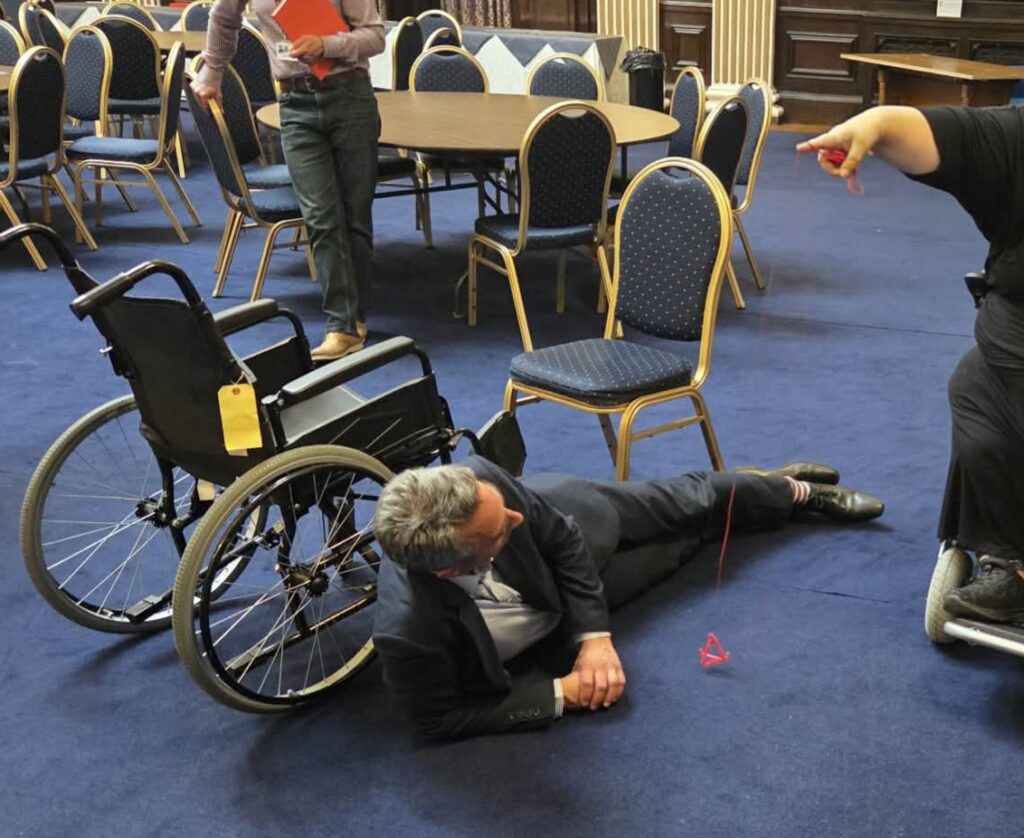 A room full of chairs and tables in the background, with a man laid on the fall in front of a wheelchair