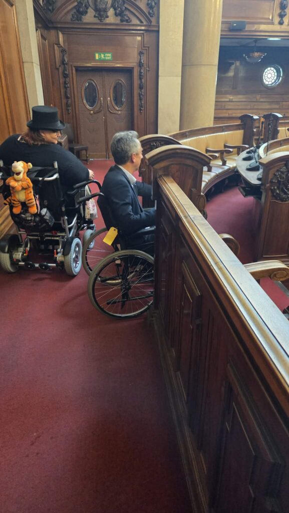 two wheelchair users on a rich burgandy coloured carpet surrounded by high wooden pew like structures