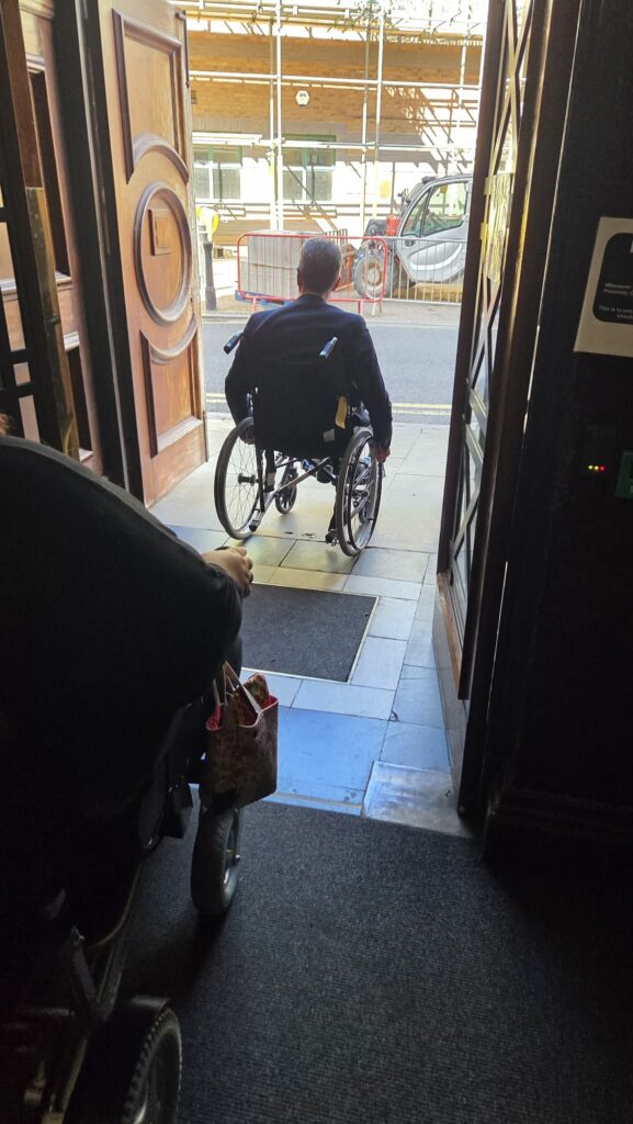 A man in a manual wheelchair leaving a dark building into a bright street beyond